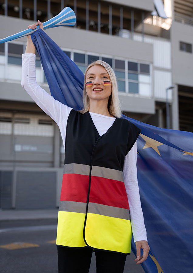 Jubelnde Erwachsene Frau steht in schwarz-rot-gelber Korntex Warnweste mit Deutschland-Flagge und Fan-Make-up vor einem Stadion